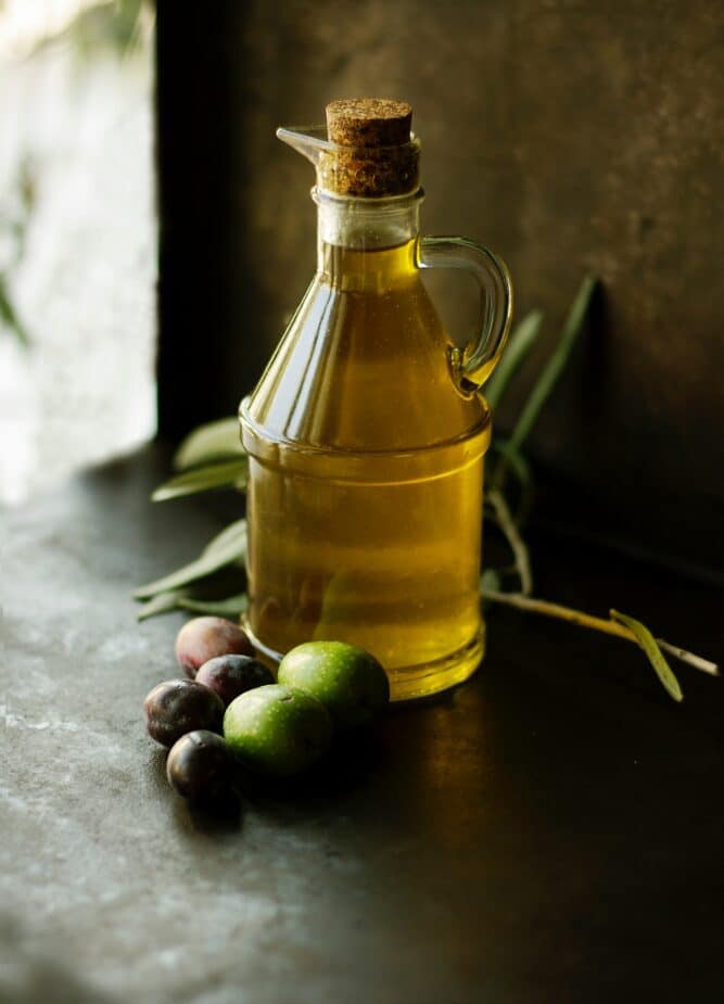 A clear glass cruet filled with olive oil on a black counter with olives and olive leaves around the base. A drizzle of these healthy oils is a great way to give your mood a boost