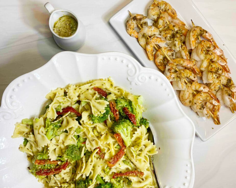 A white bowl filled with pesto pasta salad with grilled shrimp on skewers on a white platter next to it. .