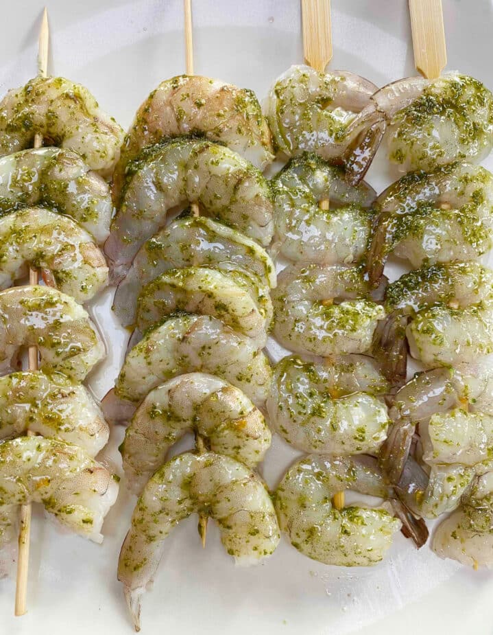 4 skewers of shrimp with pesto on a white plate before grilling