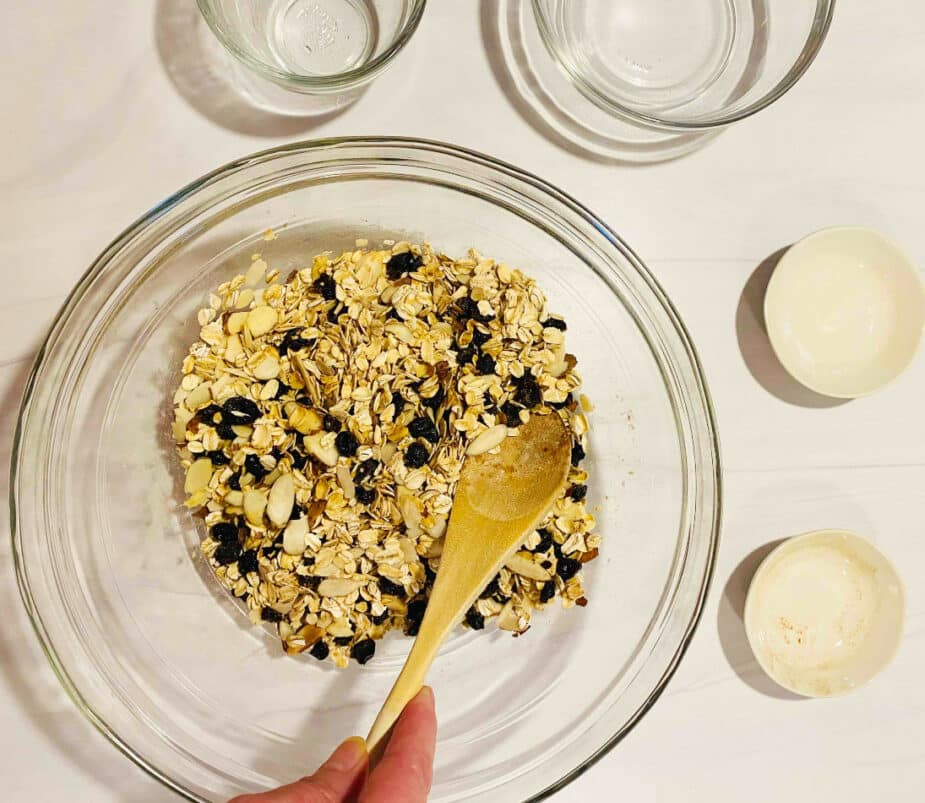 A glass bowl on a marble counter filled with dry oats, fall spices, raisins and almonds.