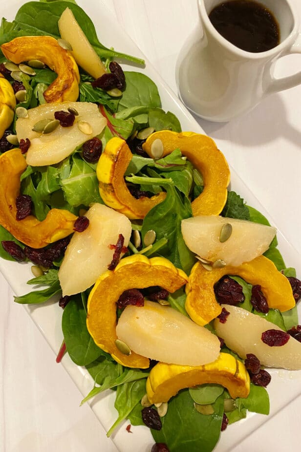 A platter of power greens topped with roasted delicata squash and pear salad. A small pitcher of balsamic vinaigrette is on the side.