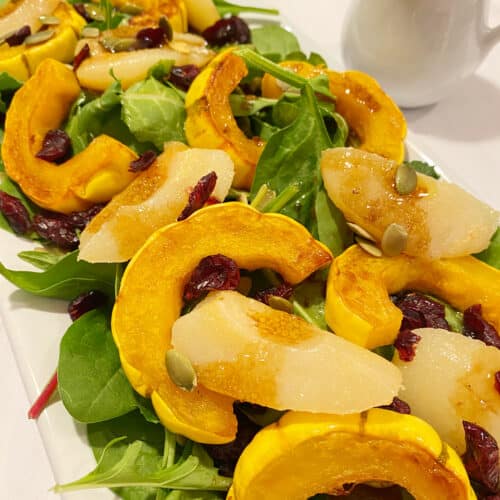 A platter of hearty power greens topped with roasted delicata squash, pears, cranberries and pumpkin seeds on a white counter.