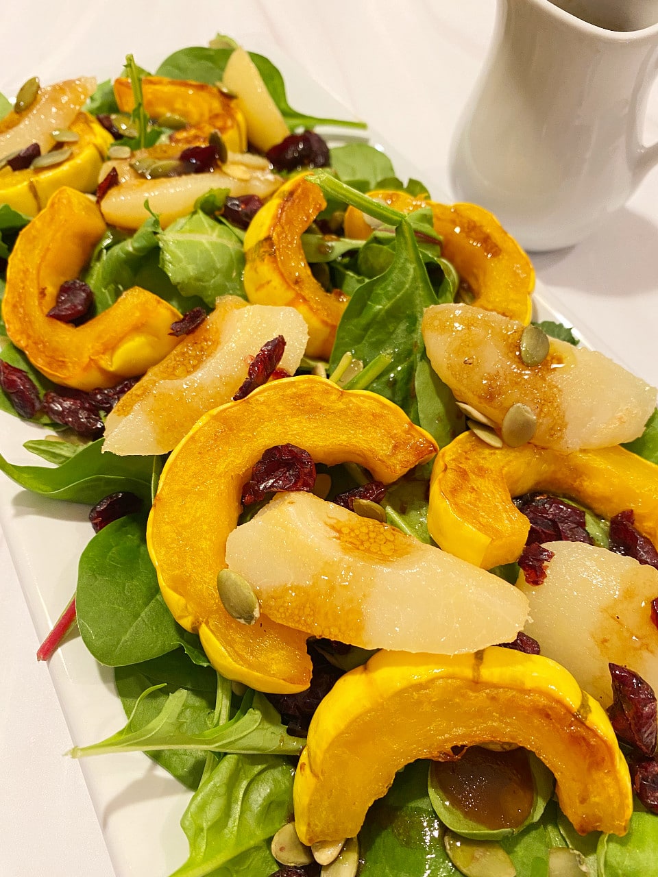 A platter of hearty power greens topped with roasted delicata squash, pears, cranberries and pumpkin seeds on a white counter.