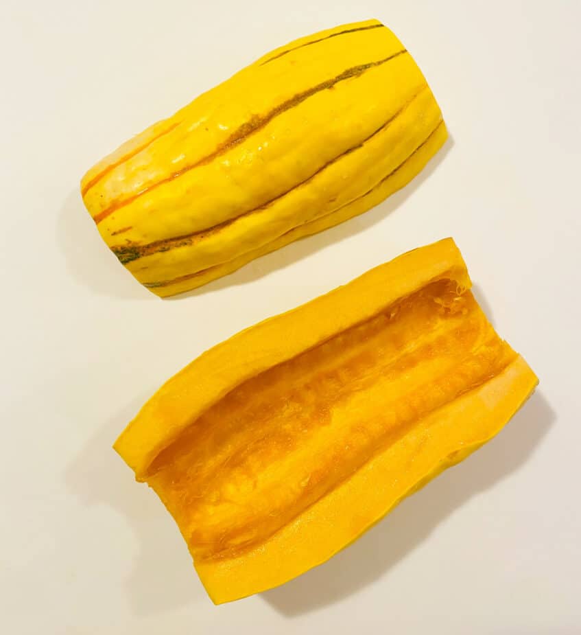 A delicata squash cut in half with the seeds scooped out sitting on a white cutting board.
