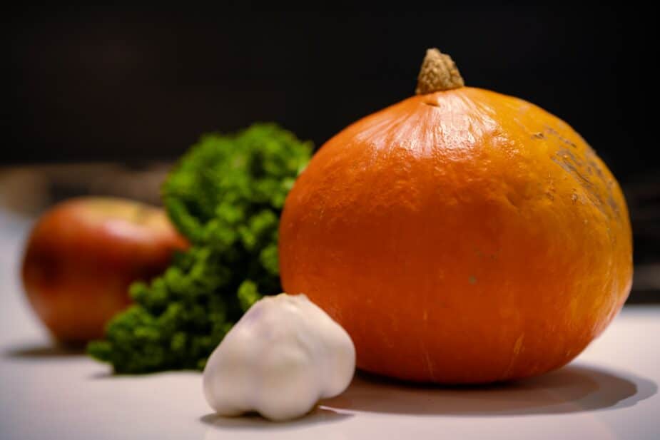 A small baking pumpkin with a bunch of garlic, parsley and an apple in the background.