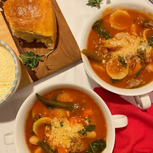 Two white bowls filled with a tomato based Minestrone soup with Italian sausage. A wooden board is on the side with grated Parmeasan cheese and dinner rolls