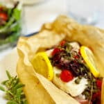 Cod with tomatoes, olives, capers and lemons in an open piece of parchment paper sitting on a white plate.