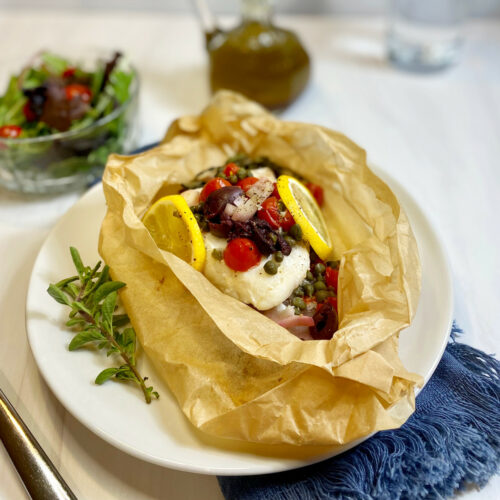 An opened parchment paper packet filled with Cod, tomatoes, olives and capers. Lemon slices are on the sides with fresh oregano. A side salad is in the background