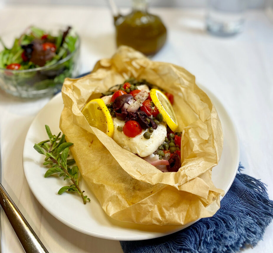 An opened parchment paper packet filled with Cod, tomatoes, olives and capers. Lemon slices are on the sides with fresh oregano. A side salad is in the background