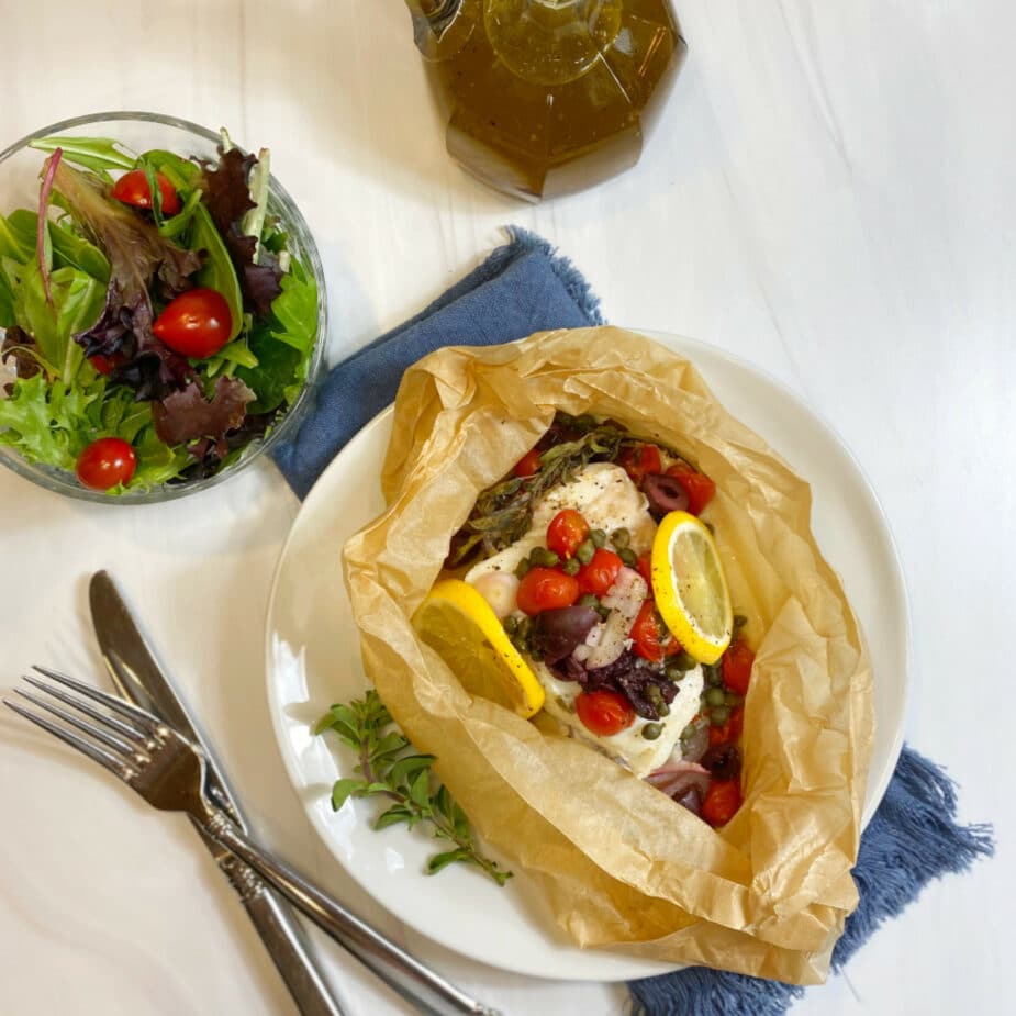 A white plate with Cod that has been baked in parchment paper with tomatoes, olives, capers and lemons.