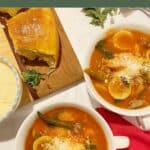 2 bowls of a rich tomato-based minestrone soup that is flavored with Italian sausage sitting on a marble counter. A wooden board with crusty rolls and a dish of grated parmeasan cheese is in the background.