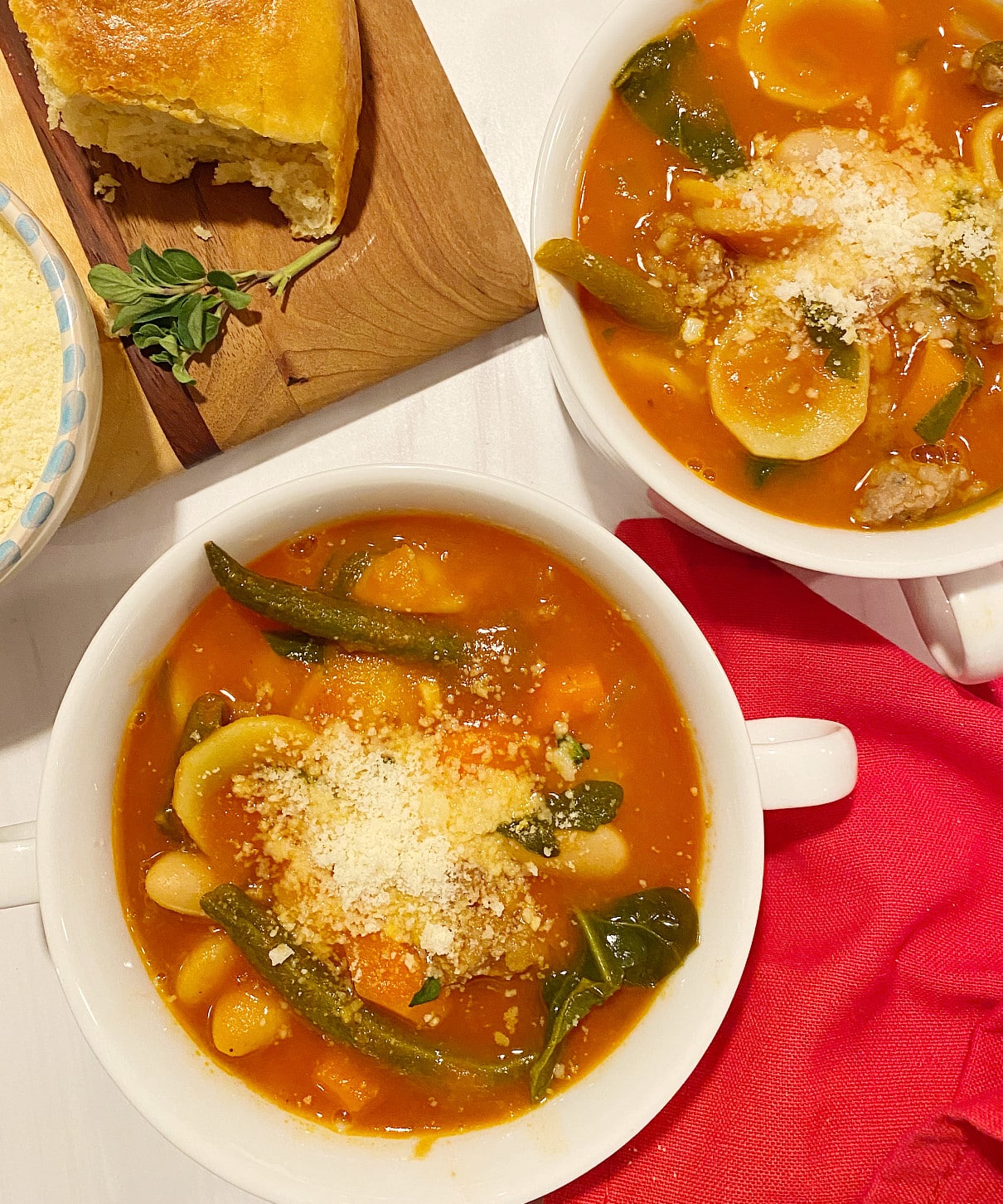 2 bowls of a rich tomato-based minestrone soup that is flavored with Italian sausage sitting on a marble counter. A wooden board with crusty rolls and a dish of grated parmeasan cheese is in the background.