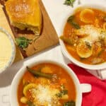 2 bowls of a rich tomato-based minestrone soup that is flavored with Italian sausage sitting on a marble counter. A wooden board with crusty rolls and a dish of grated parmeasan cheese is in the background.