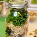 Ingredients for an ancient grains bowl packed in a mason jar on a marble counter. A colander of fresh kale, bowl of farro and chopped walnuts are in the background.