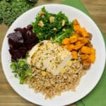 A white bowl filled with farro, roasted chicken, beets, sweetpotatoes, and kale on a dark beige wooden picnic table.