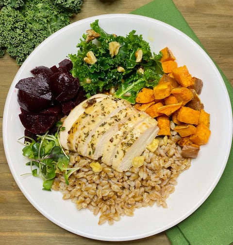 A white bowl filled with farro, roasted chicken, beets, sweetpotatoes, and kale on a dark beige wooden picnic table.