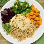 A white bowl filled with farro, roasted chicken, beets, sweetpotatoes, and kale on a dark beige wooden picnic table.