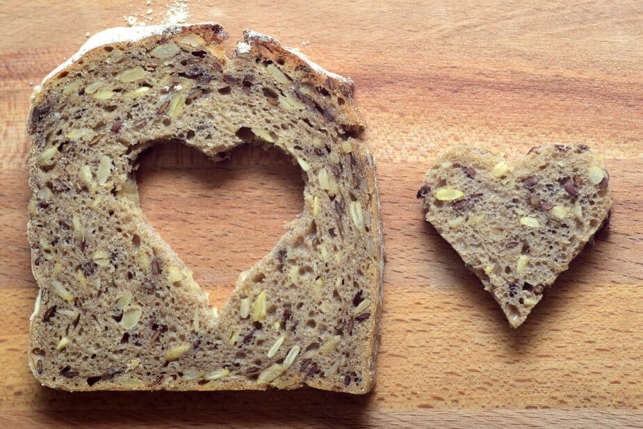 A slice of whole grain bread with a heart cut out of the center. The bread is on a wooden cutting board.