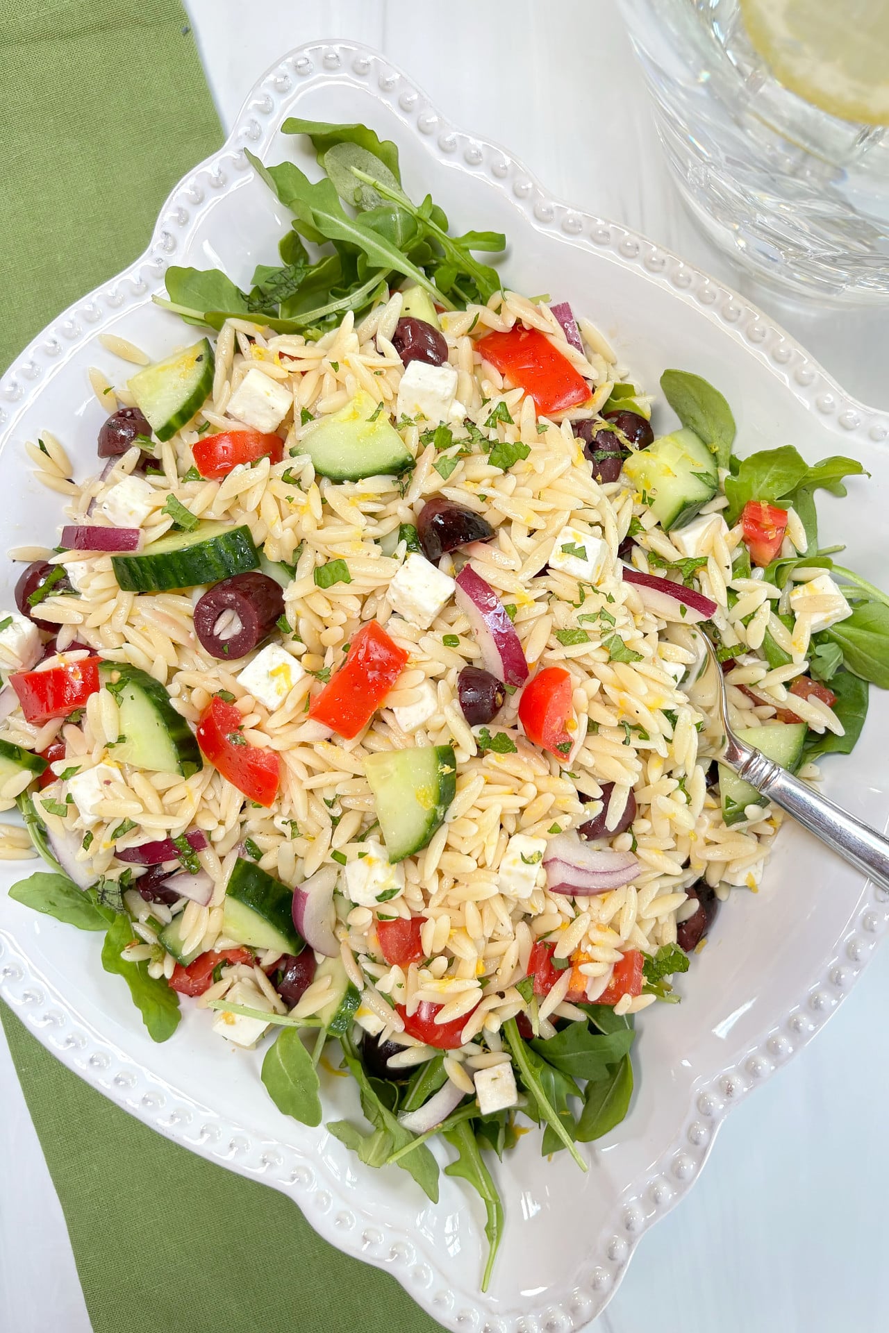 A white bowl filled with Lemon Orzo Feta Salad on a bed of arugula. It is sitting on a green cloth napkin with a pitcher of lemon water in the background.