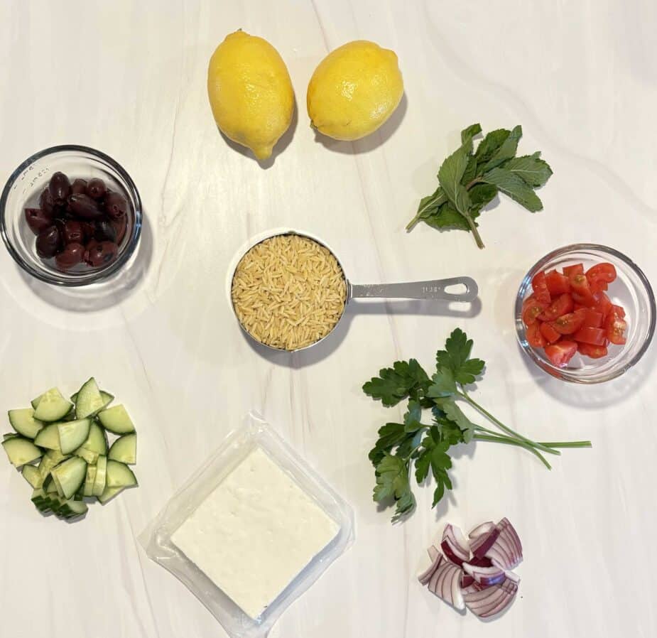 Ingredients to make a lemon orzo feta salad on a white marble counter top.