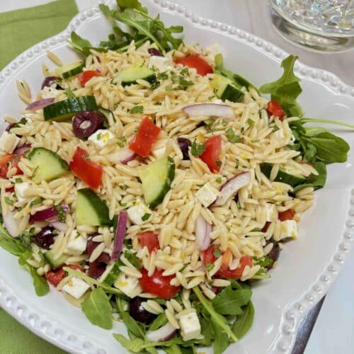 A white bowl filled with Lemon Orzo Feta Salad on a bed of arugula. It is sitting on a green cloth napkin with a pitcher of lemon water in the background.