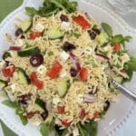 A white bowl filled with Lemon Orzo Feta Salad on a bed of arugula. It is sitting on a green cloth napkin with a pitcher of lemon water in the background.