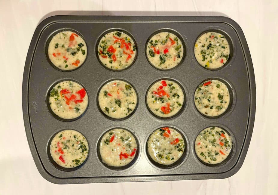 A muffin tin filled with cottage cheese and whipped egg whites, roasted red peppers and kale chips sitting on a marble counter. It is ready to go in the oven.