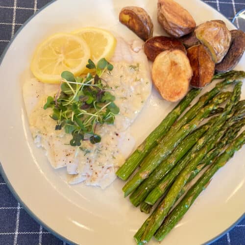 A plate on a blue placemat that has a poached flounder fillet with sauce, roasted potatoes and roasted asparagus.