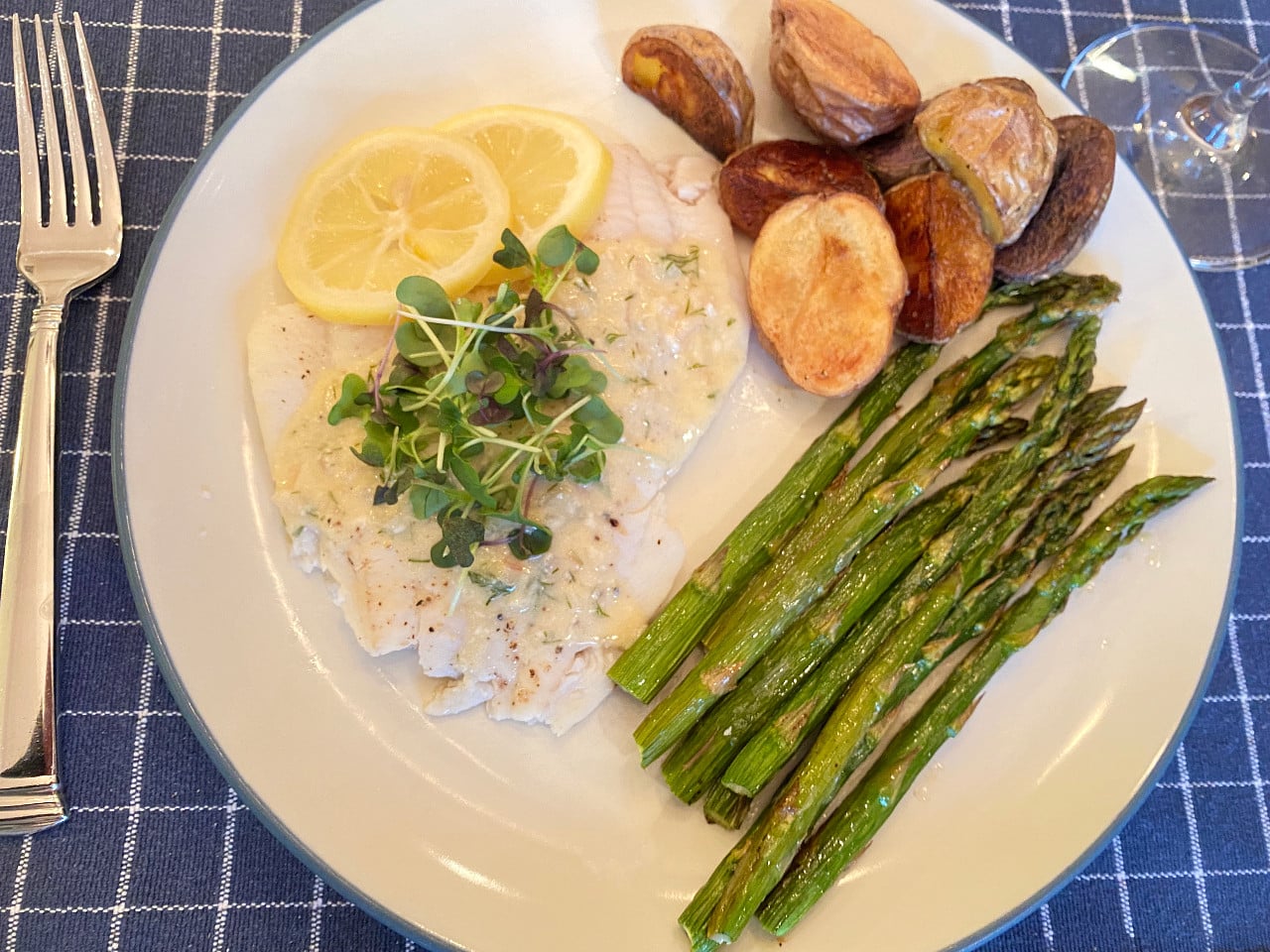 A plate on a blue placemat that has a poached flounder fillet with sauce, roasted potatoes and roasted asparagus.