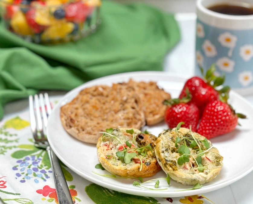 A white plate with 2 cottage cheese egg muffins, a whole grain English muffin and strawberries. A bowl of fruit salad is in the background and cup of coffee on the side.