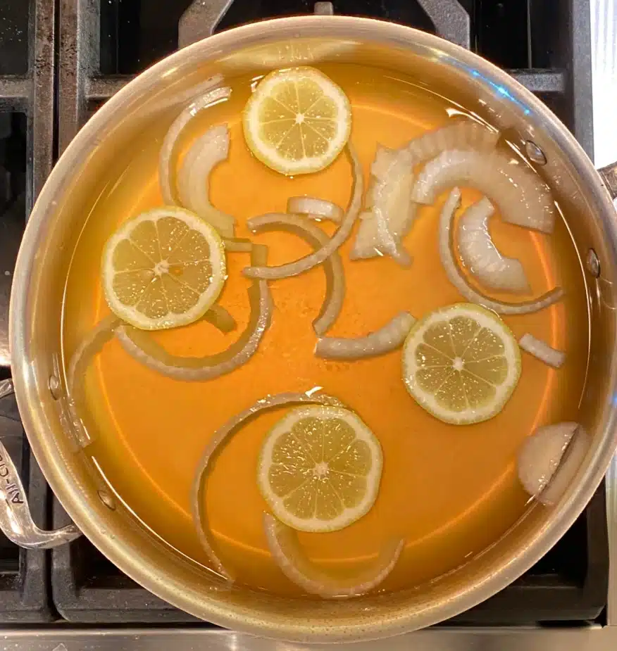 A saute pan filled with vegetable broth with onions and lemon slices dispersed throughout. This is ready to heat to poach fish in it. 