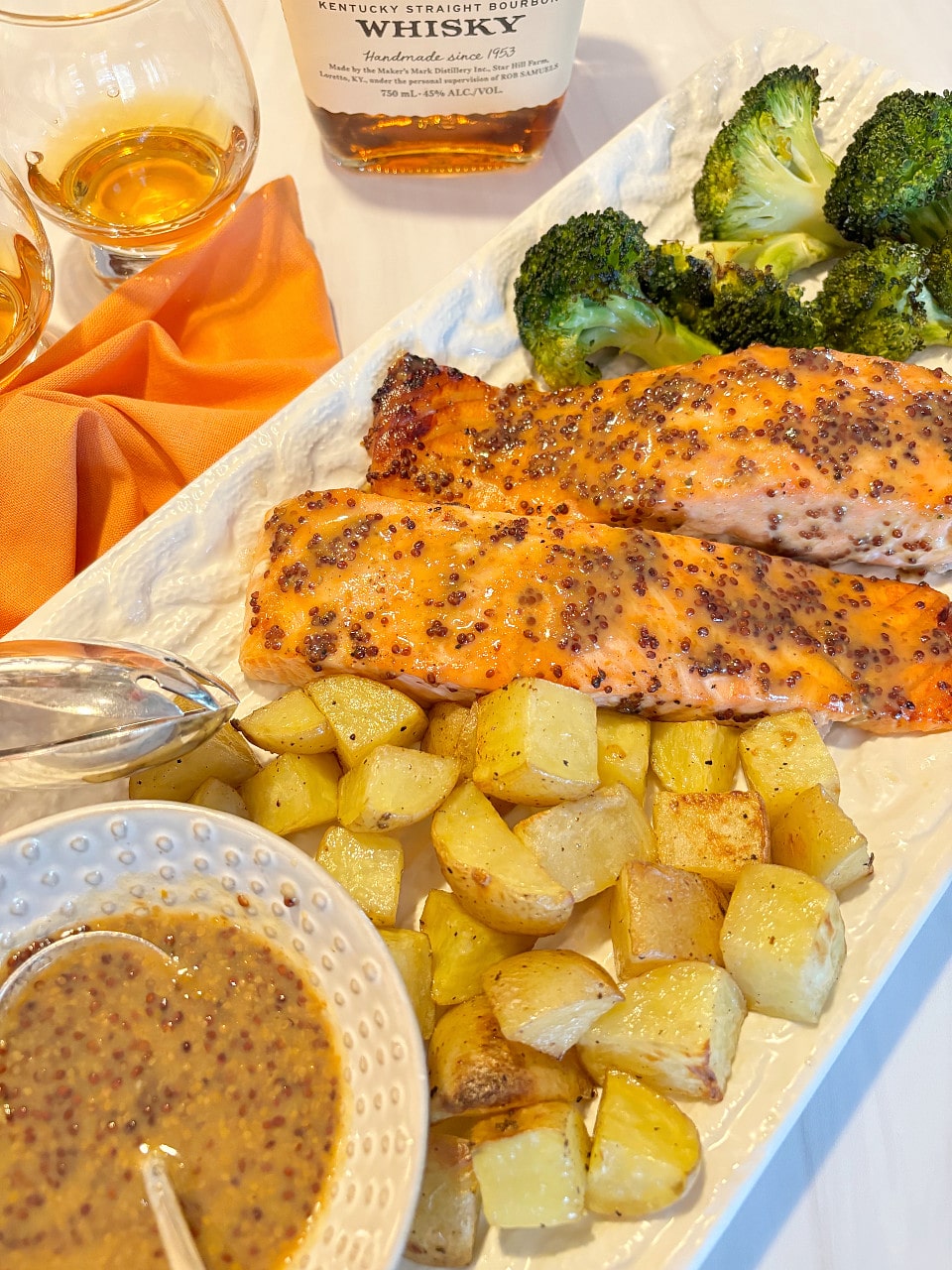 A white platter with baked cedar plank salmon in the center surrounded by roast potatoes and broccoli and extra bourbon honey mustard sauce. A bottle of bourbon is in the background.