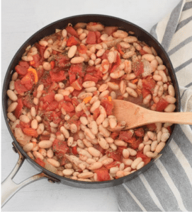 A sautepan full of cannellini beans, tomatoes and vegetables on a white background with a grey dishcloth