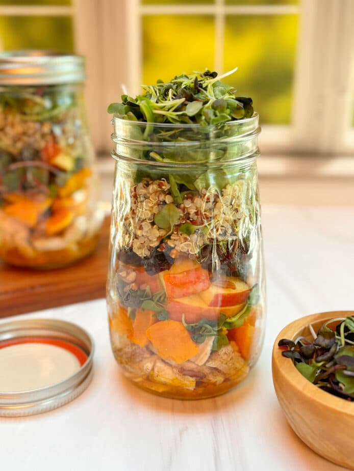 A tall mason jar filled with layers of vegetables, fruit and protein topped wit leafy microgreens. 