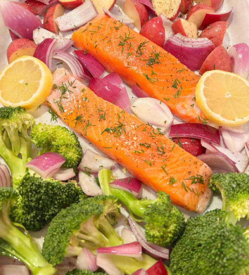 Salmon fillets surrounded by broccoli, red onions, red potatoes and lemon slices on a parchment paper lined sheet pan before roasting. 