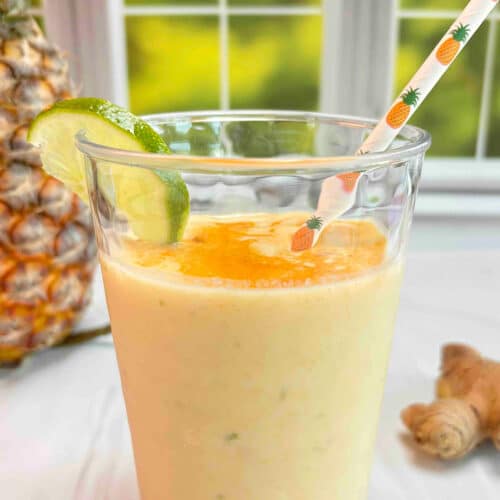 A clear glass filled with a pineapple ginger smoothie with turmeric sprinkled on top. A pineapple and piece of ginger root are in the background.