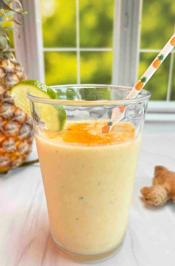 A clear glass filled with a pineapple ginger smoothie with turmeric sprinkled on top. A pineapple and piece of ginger root are in the background.