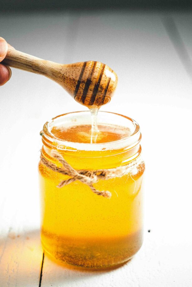 A clear jar of honey with a honey dipper drizzling honey into the jar