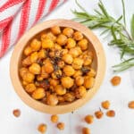 A wooden bowl filled with roasted rosemary chickpeas on a white counter with a fresh rosemary branch and red and white dishtowel in the background.