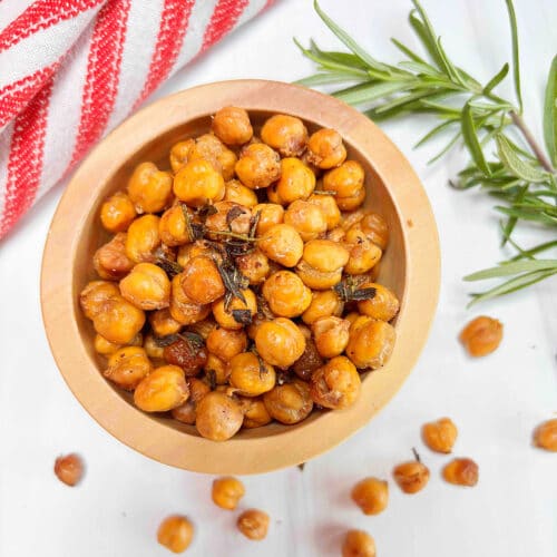 A wooden bowl filled with roasted rosemary chickpeas on a white counter with a fresh rosemary branch and red and white dishtowel in the background.