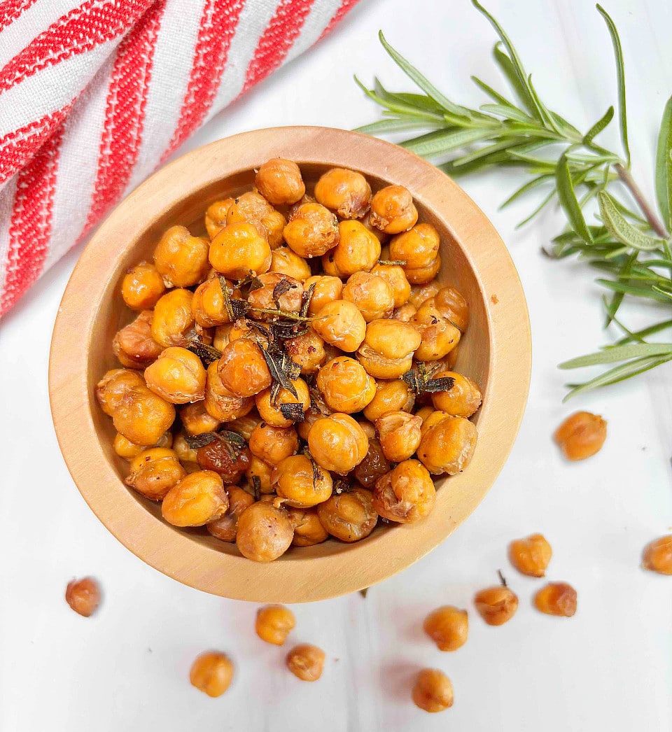 A wooden bowl filled with roasted rosemary chickpeas on a white counter with a fresh rosemary branch and red and white dishtowel in the background.