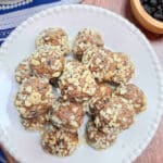 A plate of almond blueberry energy bites on a wood table with a bowl of dried blueberries and a blue dish towel in the background