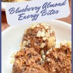 A plate with a blueberry almond energy bite cut open to show the texture next to an unopened ball. A plate of the energy bites are in the background.