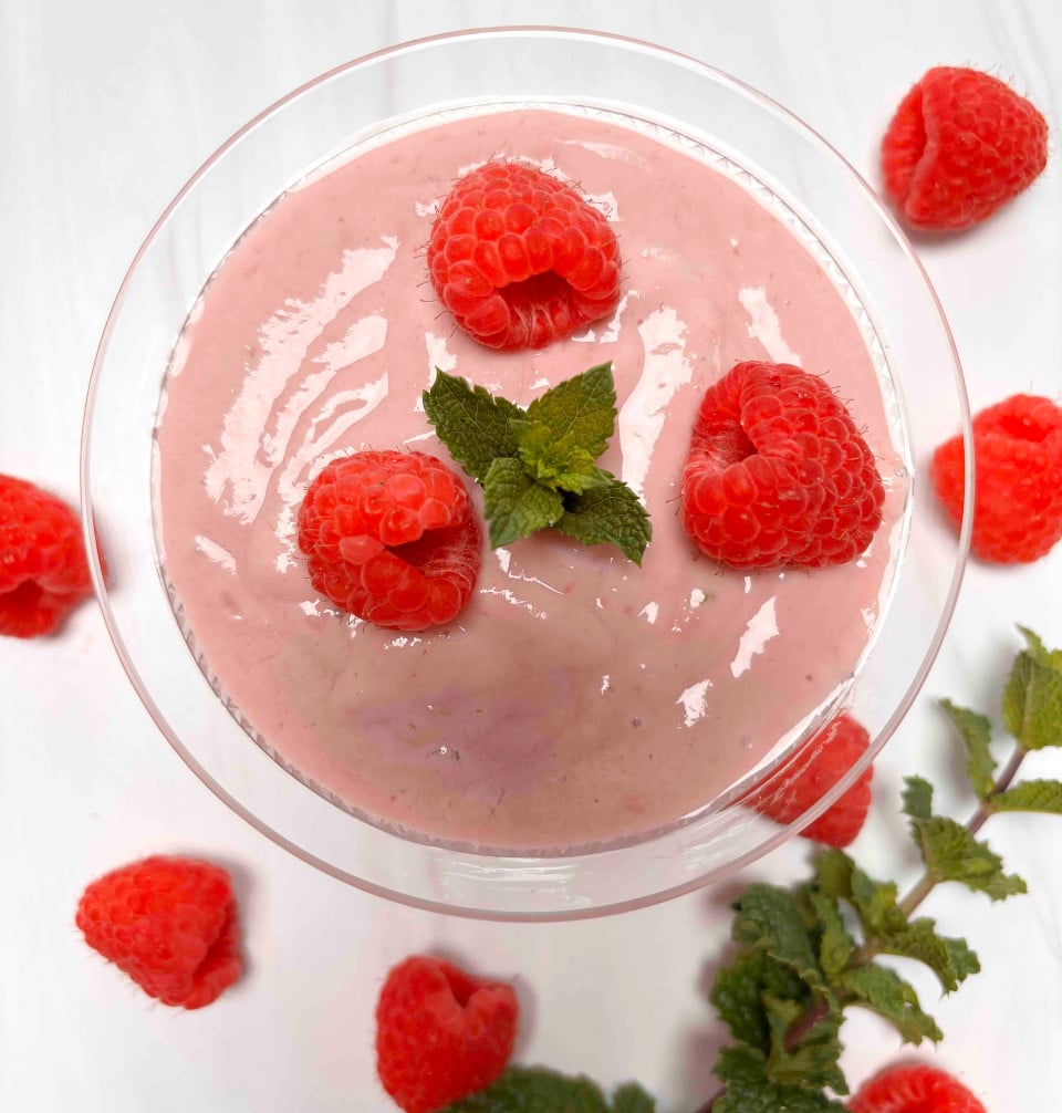 Raspberry Silken Tofu Mousse in a glass cup garnished with fresh raspberries and mint on a white counter.