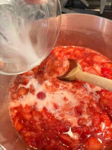 Adding cornstarch mixed with water to a saucepan filled with partially cooked strawberries and sugar.