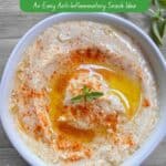 A white bowl filled with creamy hummus garnished with smoked paprika and fresh oregano sitting on a wooden picnic table.