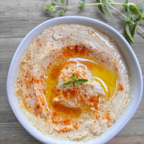 A white bowl filled with creamy hummus on a wood table. The hummus is garnished with a sprinkle of smoked paprika and a sprig of fresh oregano.