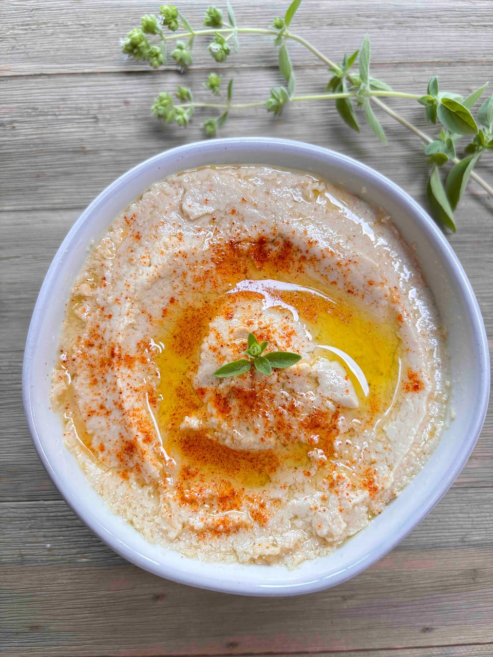 A white bowl filled with creamy hummus on a wood table. The hummus is garnished with a sprinkle of smoked paprika and a sprig of fresh oregano.