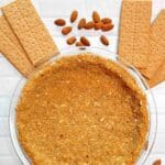 A graham cracker pie crust in a glass pie pan surrounded by graham crackers and almonds.