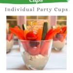 A glass cup with the bottom filled with hummus and a variety of veggie sticks and crackers dipped in the hummus and poking out the top of the glass. Additional cups are in the background.